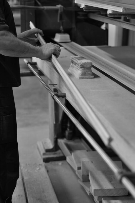Tehnician productie verifică un profil lung din lemn pe masa de lucru, alături de bacuri de fixare și materiale pregătite pentru prelucrare